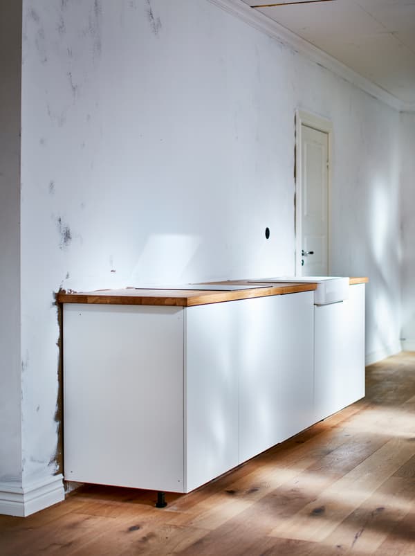 Dapur kecil sebaris berwarna putih dengan permukaan atas kerja kayu di dalam bilik yang kosong tanpanya dengan lantai kayu dan dinding berwarna putih.