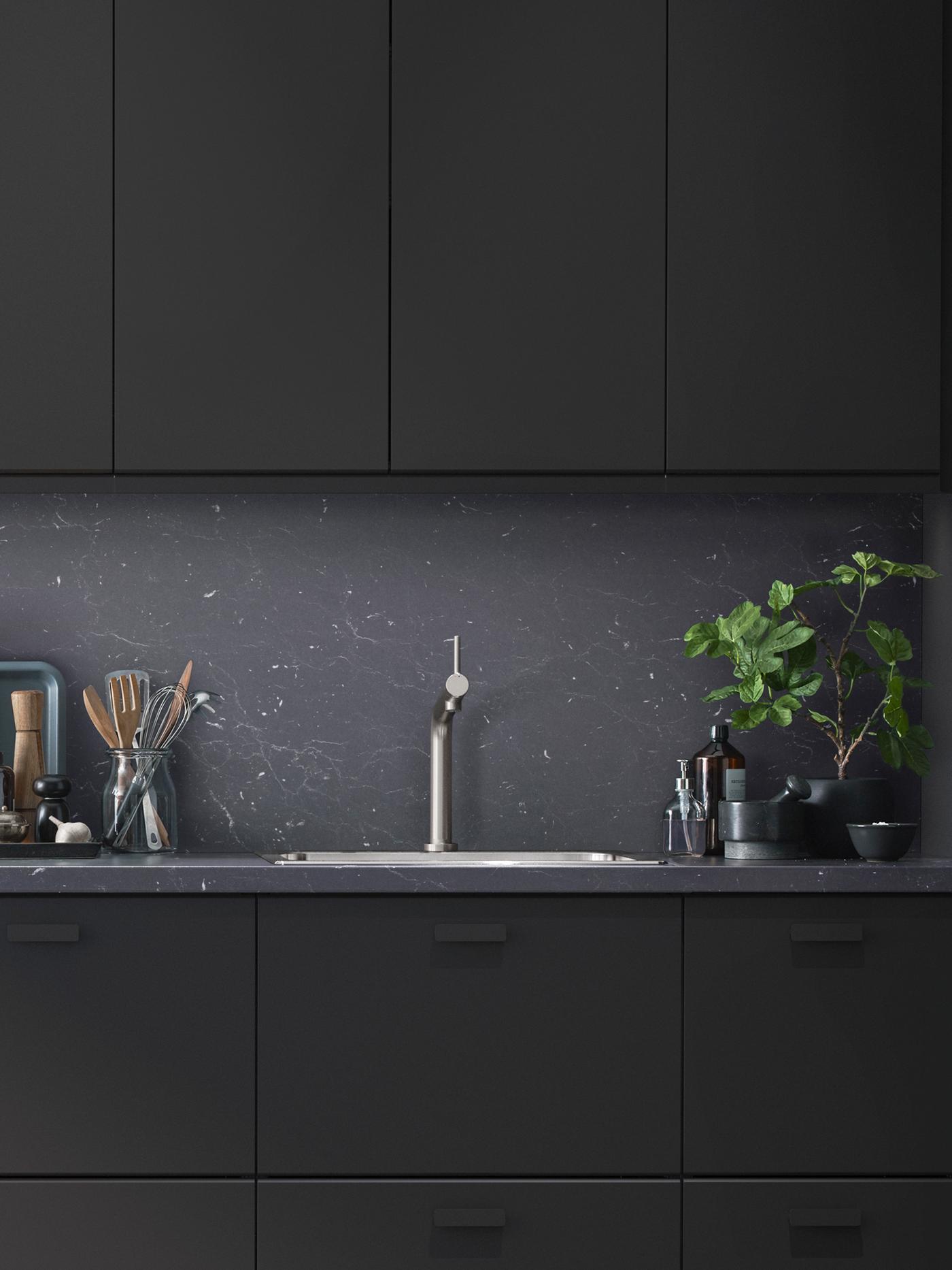 Dapur moden dengan bahagian hadapan KUNGSBACKA berwarna hitam dan perkakas pada satu sisi sink dan tumbuhan besar di satu sisi lagi.