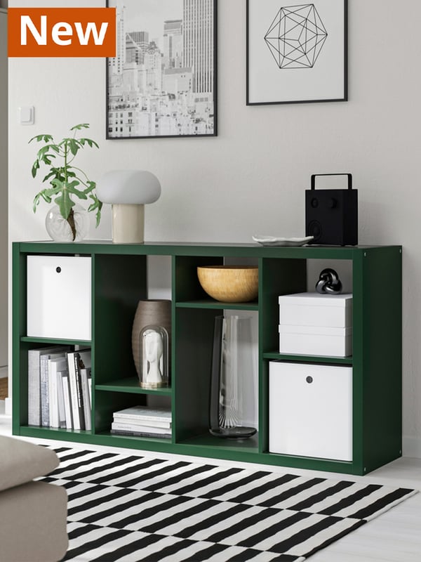 Dark green KALLAX shelf lying horizontally across in front of a white-painted wall. Some white DRÖNA boxes are inserted within the KALLAX on top of some books and decorative items.