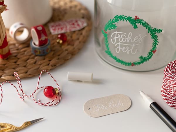 Das bemalte Glas wird mit Geschenkanhängern, Geschenkbändern und Dekokugeln dekoriert.
