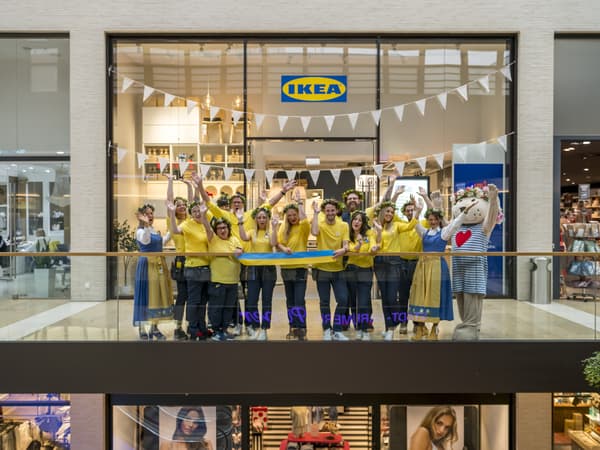 Das Bild zeigt die Eröffnung eines IKEA Planungsstudios in einem Einkaufszentrum in Rheine. Ein fröhliches IKEA Team in gelben Hemden vor dem Studio und jubelt, während sie ein blau-gelbes Band halten. Einige Teammitglieder tragen Blumenkränze auf dem Kopf, und links im Bild steht ein Maskottchen im IKEA Stil. Die Dekoration umfasst weiße Wimpelketten, und im Hintergrund ist das IKEA Logo über dem Eingang des Planungsstudios zu sehen.