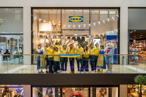 Das Bild zeigt die Eröffnung eines IKEA Planungsstudios in einem Einkaufszentrum in Rheine. Ein fröhliches IKEA Team in gelben Hemden vor dem Studio und jubelt, während sie ein blau-gelbes Band halten. Einige Teammitglieder tragen Blumenkränze auf dem Kopf, und links im Bild steht ein Maskottchen im IKEA Stil. Die Dekoration umfasst weiße Wimpelketten, und im Hintergrund ist das IKEA Logo über dem Eingang des Planungsstudios zu sehen.