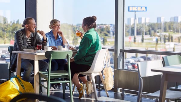 Das Bild zeigt drei Personen, die in einem IKEA Restaurant an einem Tisch sitzen und sich unterhalten. Im Hintergrund ist durch die Fenster ist das IKEA Logo zu sehen. Die Atmosphäre wirkt entspannt und freundlich. Eine der Personen hält ein Getränk in der Hand, während eine andere ein Gebäckstück genießt. Auf dem Boden neben dem Tisch liegt eine große gelbe IKEA Einkaufstasche. Der helle und moderne Raum bietet einen angenehmen Blick nach draußen auf die Umgebung, die von Bäumen und einem Parkplatz geprägt ist.