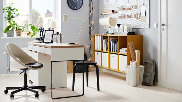 Das Bild zeigt ein helles und modern eingerichtetes Arbeitszimmer. Ein weißer Schreibtisch mit Holzplatte steht im Vordergrund, darauf befinden sich eine Lampe, Stiftehalter und ein Tablet. Ein ergonomischer Bürostuhl sowie ein kleiner schwarzer Hocker sind ebenfalls zu sehen. Im Hintergrund gibt es ein gelbes Regal mit weißen Aufbewahrungsboxen und verschiedenen Büchern. An der Wand hängen Dekorationen und Lichterketten, die dem Raum eine gemütliche Atmosphäre verleihen. Eine große Pflanze steht auf der Fensterbank und bringt etwas Grün in den Raum.