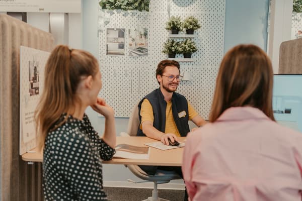 Das Bild zeigt eine Beratungsszene in einem IKEA Planungsstudio. Ein lächelnder IKEA Mitarbeiter sitzt an einem Schreibtisch vor einem Computer und spricht mit zwei Kundinnen. Im Hintergrund ist eine Wand mit dekorativen Pflanzen und Küchenbildern zu sehen. Der Mitarbeiter trägt eine IKEA Weste und arbeitet an der Planung eines Raumes, während die Kundinnen aufmerksam zuhören.