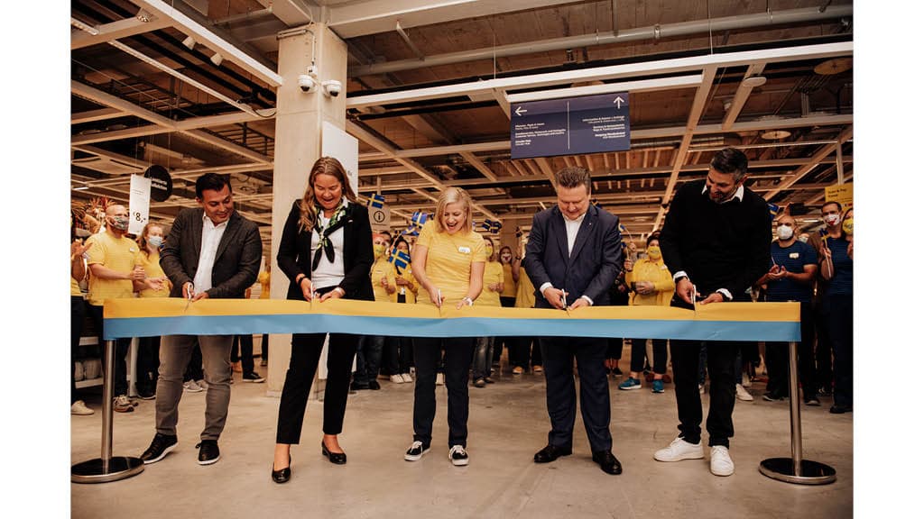 Das Bild zeigt eine feierliche Eröffnung mit fünf Personen, die gemeinsam ein blau-gelbes Band durchschneiden. Im Hintergrund stehen zahlreiche Mitarbeitende in gelben Hemden