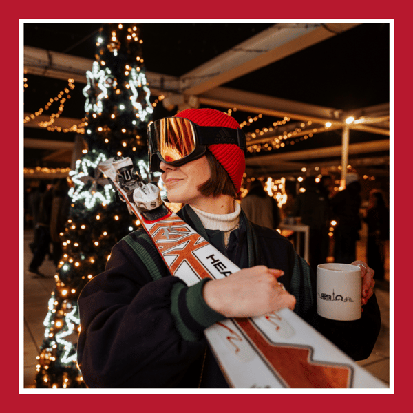 Das Bild zeigt eine Frau mit Ski über den Schultern und einer Tasse in der Hand mit einem Weihnachtsbaum im Hintergrund.