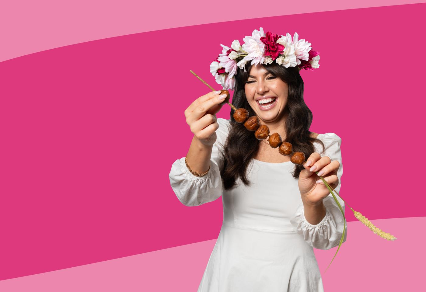 Das Bild zeigt eine Person in einem weißen Kleid mit Blumenkranz auf dem Kopf, die eine Holzspießkette mit Fleischbällchen in die Kamera hält. Der Hintergrund ist in kräftigen Pinktönen gestaltet.