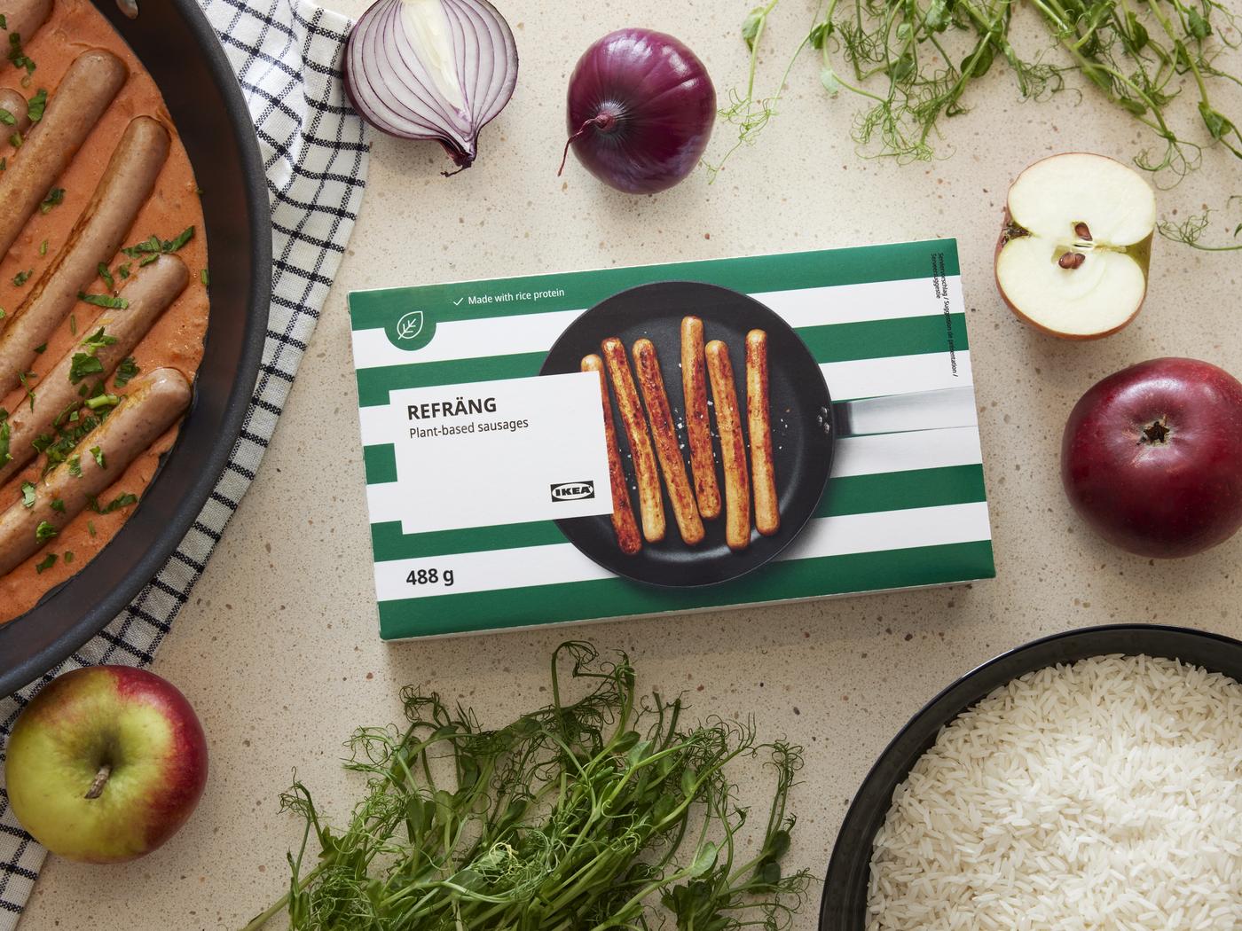 Das Bild zeigt eine Verpackung von IKEA mit der Aufschrift „REFRÄNG – Plant-based sausages“, also pflanzenbasierte Würstchen. Die Verpackung hat grüne und weiße Streifen. Auf der Verpackung ist eine Pfanne mit fünf Würstchen abgebildet. Rechts daneben liegen zwei rote und ein grüner Apfel, sowie eine halbe Zwiebel. Unten rechts befindet sich eine Schüssel mit Reis. Links im Bild ist eine Pfanne mit pflanzlichen Würstchen, die in einer Soße garen, zu sehen. Daneben liegt ein Küchentuch. Oben und unten im Bild sind zudem frische Kräuter und weitere Zutaten wie eine aufgeschnittene rote Zwiebel platziert.