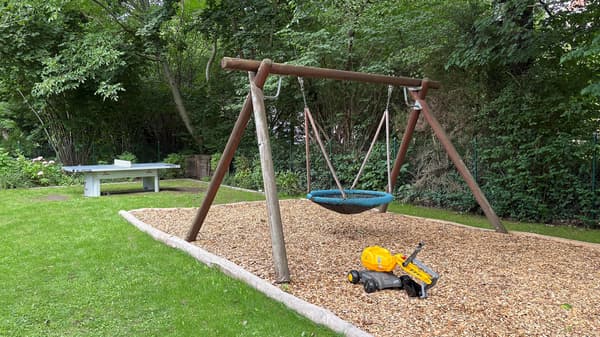 Das Bild zeigt einen Gartenbereich mit einer großen Nestschaukel aus Holz. Die Schaukel hängt an einem stabilen Holzgestell, der Boden darunter ist mit Holzhackschnitzeln bedeckt, um Stürze abzufedern. Im Vordergrund liegt ein gelbes Spielzeug-Baufahrzeug auf dem Boden. Im Hintergrund steht eine fest installierte Tischtennisplatte auf einer Rasenfläche. Der Garten ist von Bäumen und viel Grün umgeben.