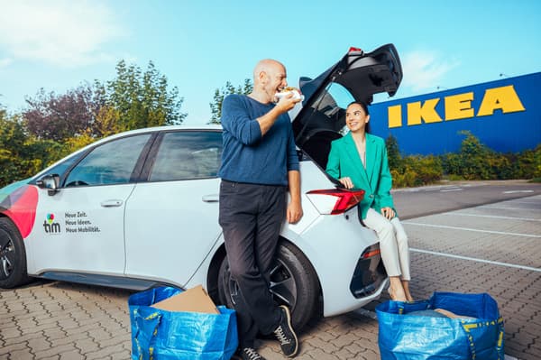 Das Bild zeigt zwei Personen auf dem IKEA-Parkplatz mit einem weißen Mietauto. Sie haben offenbar eingekauft, denn rund um das Auto stehen mehrere volle IKEA-Taschen. 