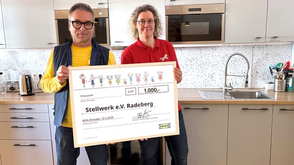 Das Bild zeigt zwei Personen, die einen großen symbolischen Scheck von IKEA Dresden über 1.000 Euro präsentieren. Die Szene vermittelt den Moment einer Spendenübergabe in einem freundlich eingerichteten Raum.