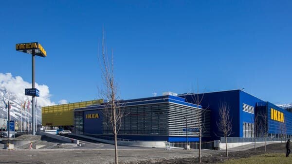 Das Eingangsportal von IKEA Innsbruck bei strahlendem Sonnenschein mit blauem Himmel und wehenden Flaggen.