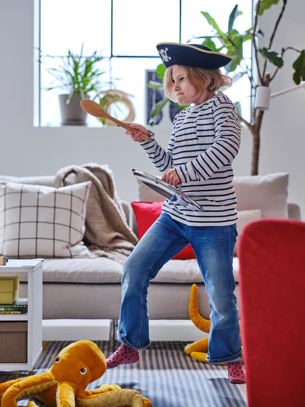 Davanti a un divano SÖDERHAMN beige, un bambino con cappello da pirata simula un duello alla spada impugnando un cucchiaio in legno RÖRT e un coperchio per casseruola - IKEA.
