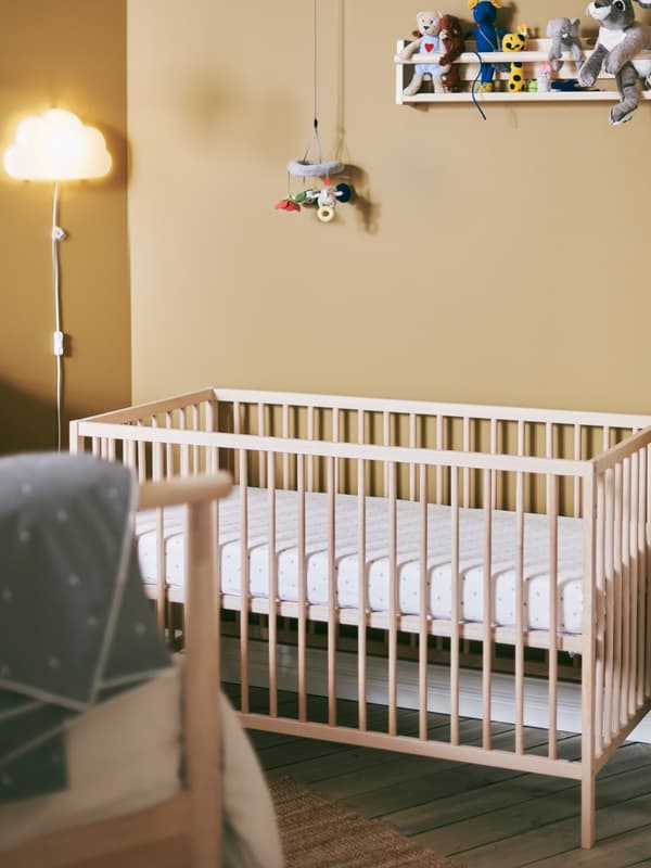 Davanti a una parete marrone, un lettino SNIGLAR con materasso rivestito da lenzuolo con angoli LENAST bianco; accanto al lettino, una lampada da parete a LED UPPLYST bianca a forma di nuvola - IKEA.
