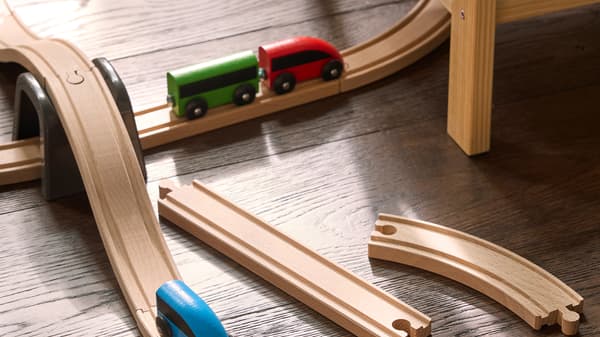 De foto toont een houten speelgoedtreinbaan met een rood-groene speelgoedtrein en extra railstukken op de vloer.