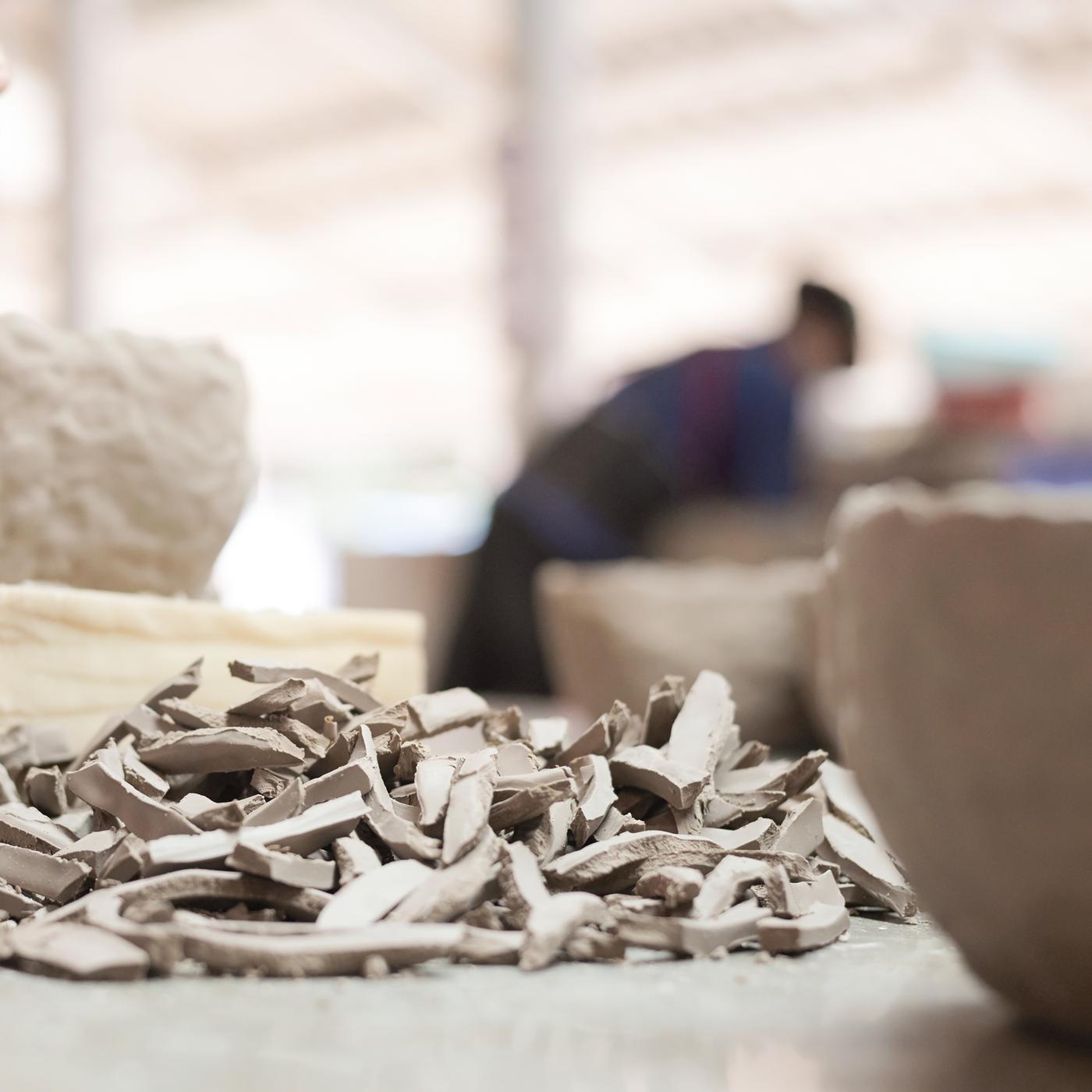 De petits morceaux de poterie brisés sont posés sur une table avec des céramiques entières plus grandes et un artisan en arrière-plan.