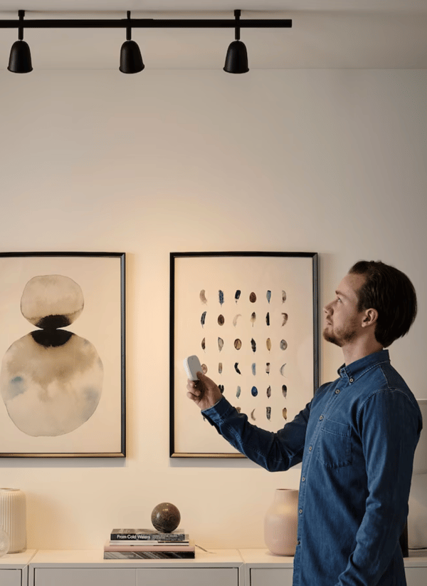Debout dans son séjour, un homme tient une télécommande pour éclairage connecté IKEA. 