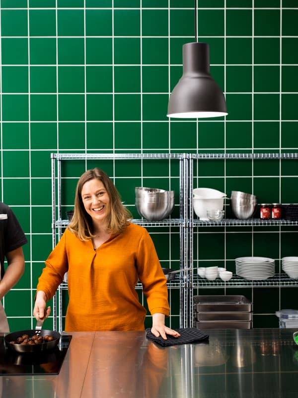 Debout dans une cuisine avec des meubles en acier inoxydable et des carreaux verts sur les murs, une femme tient une poêle contenant des boulettes végétales.