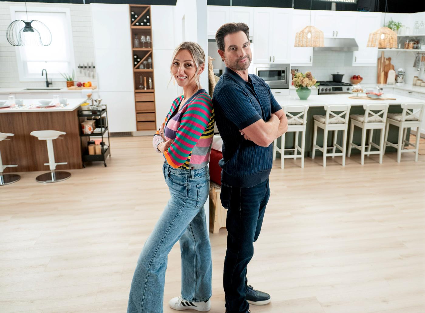Debra Salmoni, à gauche, debout dos à dos avec Scott McGillivray. Tous deux ont les bras croisés et sourient devant deux cuisines modernes. 