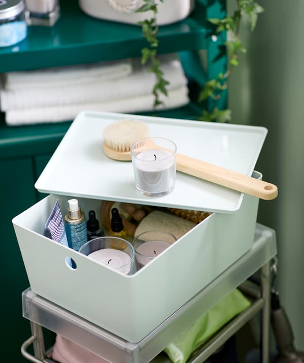 Décor de salle de bain avec un chariot rempli d’accessoires. Dessus se trouve une boîte KUGGIS contenant un kit bien-être: des huiles, des brosses, des bougies SINNLIG.