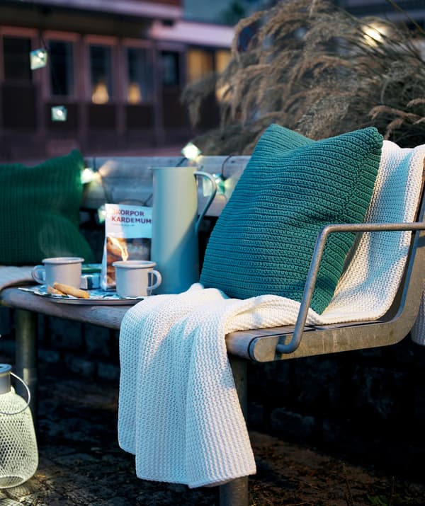 Décor de soirée en ville avec un banc public équipé d’un plateau à café, de coussins, d’un plaid, de lampes décoratives et d’une lanterne SOLVINDEN.