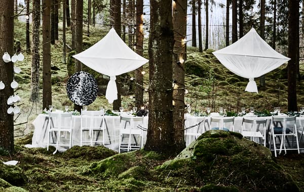 Décor de table pour une fête de mariage en forêt, avec chaises blanches, nappes en voilages et grandes décorations centrales suspendues à la manière de lustres