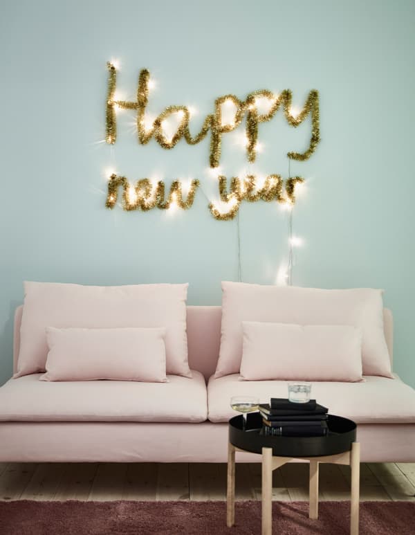 Decoración de parede para Aninovo creada cunha grilanda de luces LED con batería SÄRDAL e unha grilanda típica do Nadal dourada sobre un sofá rosa.