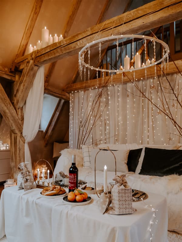 Décoration bohème en intérieur avec une table ornée d'une nappe blanche, un lustre, beaucoup de guirlandes lumineuses contre le mur du fond et des bougies posées sur les poutres en bois.