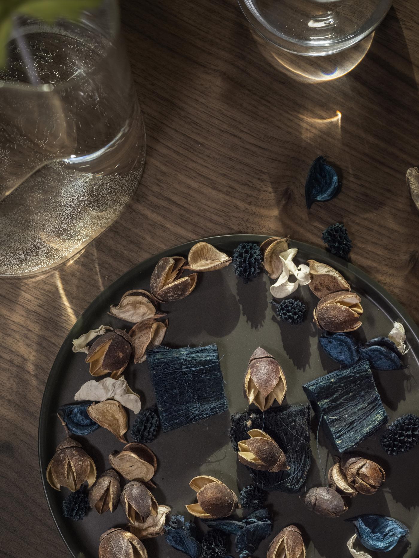 Decorative plate with dried potpourri and fabric pieces on a wooden surface, surrounded by clear glassware.