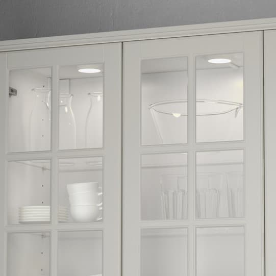 Dei faretti MITTLED bianchi, montati dentro un mobile da cucina con le ante a vetro, illuminano gli oggetti in vetro all'interno.
