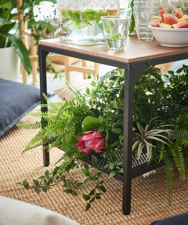 Delle piante articiali FEJKA con vaso sotto un tavolino FJÄLLBO. Sopra il tavolino diversi bicchieri e stoviglie di vetro e sotto un tappeto LOHALS.