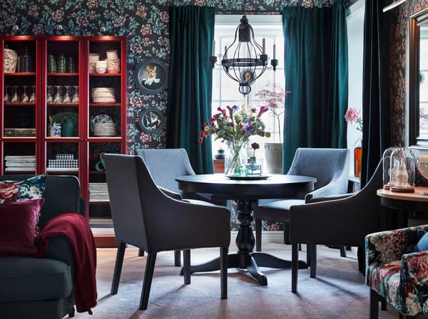 Der IKEA INGATORP Ausziehtisch in Schwarz und SAKARIAS Armlehnstühle in Schwarz mit Bezug „Sporda“ in Dunkelgrau  mit geblümten Kissen unterstreichen den klassischen Look des Essbereichs mit seiner altmodischen Blumentapete und den dunkelgrünen Gardinen aus Samt.