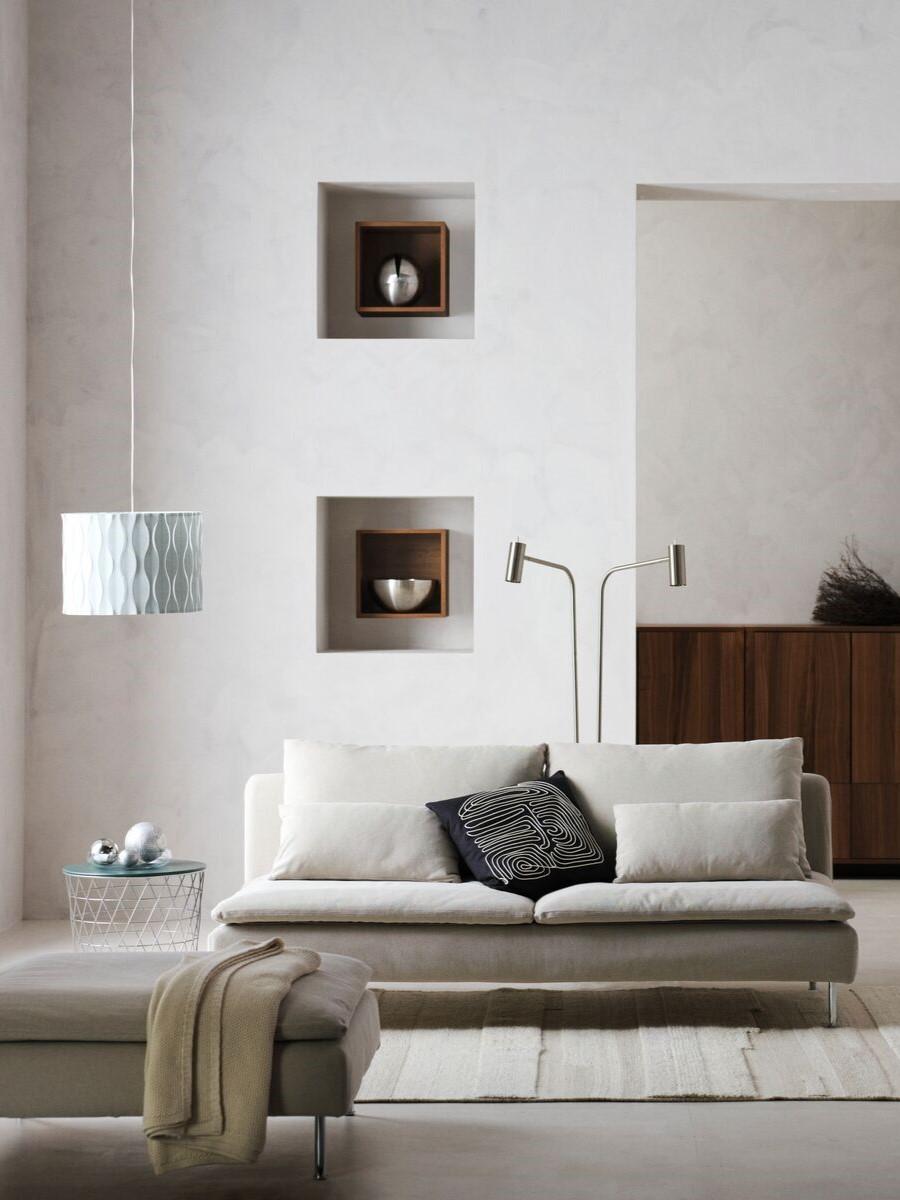 Der RAum hat einen modernen Scandi Stil als einrichtung. Es hat eine weiße Wand mit Bilder an der Seite und einem hellen Sofa davor. An der Seite steht ein dunkler Stuhl.