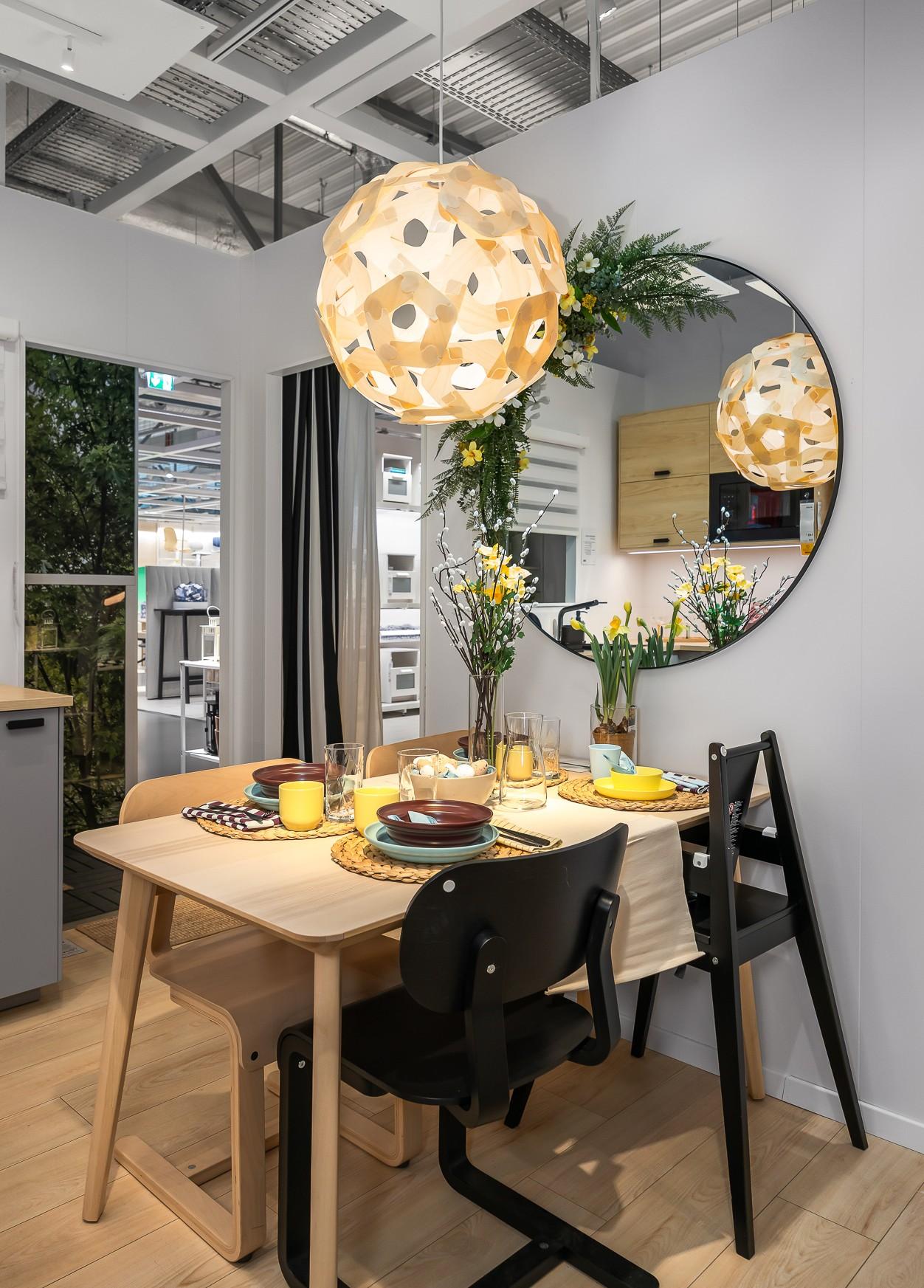 Деревянный обеденный стол, украшенный к Пасхе многослойной посудой, свечами и натуральными элементами, установленный под круглой подвесной лампой и большим зеркалом.