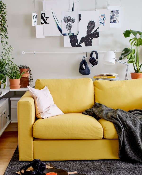 Derrière le canapé jaune, deux barres servent de support à des œuvres d’art et à un petit panier suspendu. Une plante en pot et une lampe sont installées sur une tablette. 