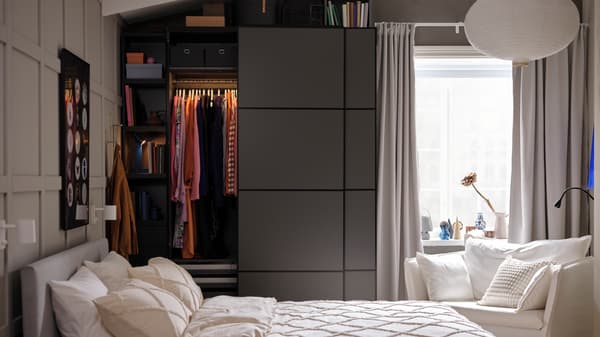 Derrière un lit, une armoire PAX/MEHAMN gris foncé dont la porte coulissante gauche ouverte laisse voir des vêtements suspendus à une tringle.