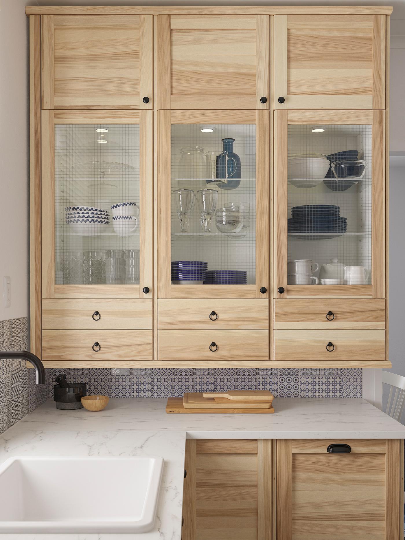 Des armoires murales avec des portes en frêne et des portes vitrées TORHAMN servant à ranger de la vaisselle au-dessus d’un comptoir blanc à effet de marbre.