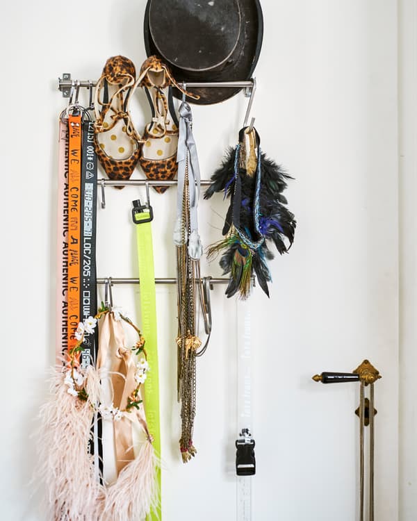 Des barres murales métalliques de cuisine sur une porte blanche. Elles accueillent des accessoires dont un haut-de-forme, des escarpins à motif léopard, des colliers et des tours de cou.