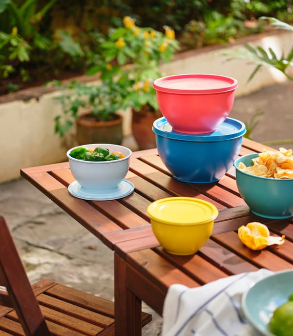 Des bols avec couvercles colorés posés sur une table en bois. On aperçoit des plantes à l’arrière-plan.