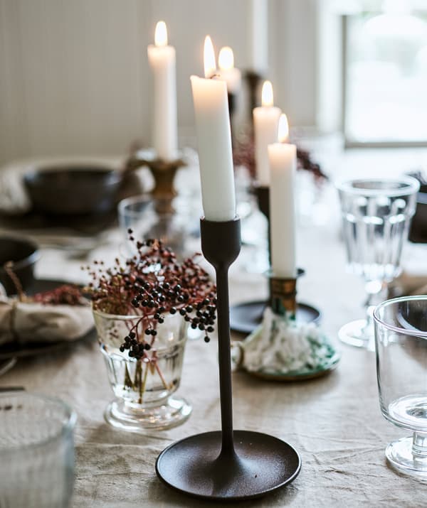 Des bougies allumées dans un mélange de bougeoirs noirs et en laiton sur une table avec une nappe en lin, des verres et des baies dans un vase.
