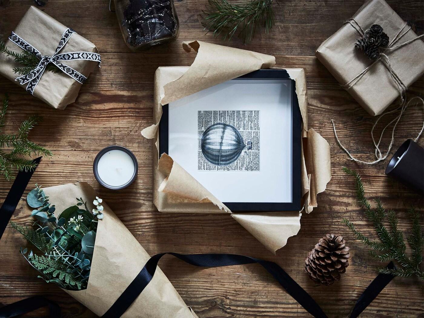 Des cadeaux partiellement emballés incluant un cadre photo, un bouquet de fleurs et une chandelle sur un plancher de bois rustique.