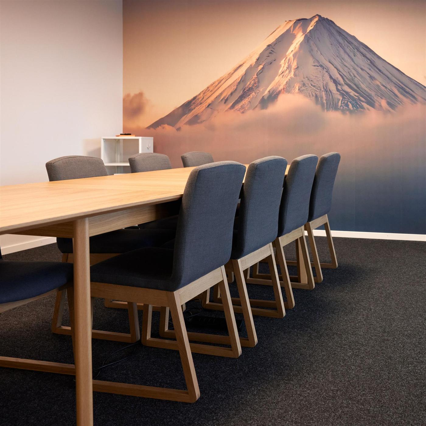 Des chaises en bois moelleuses sont disposées autour d’une grande table en bois dans une salle de réunion dont le mur du fond est décoré d’un papier peint panoramique représentant une montagne