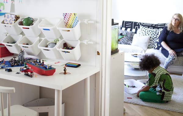 Des contenants sont suspendus à un rail mural pour créer un espace de rangement de jouets dans la chambre d’un enfant. Mère et fils sont assis à l’arrière-plan.