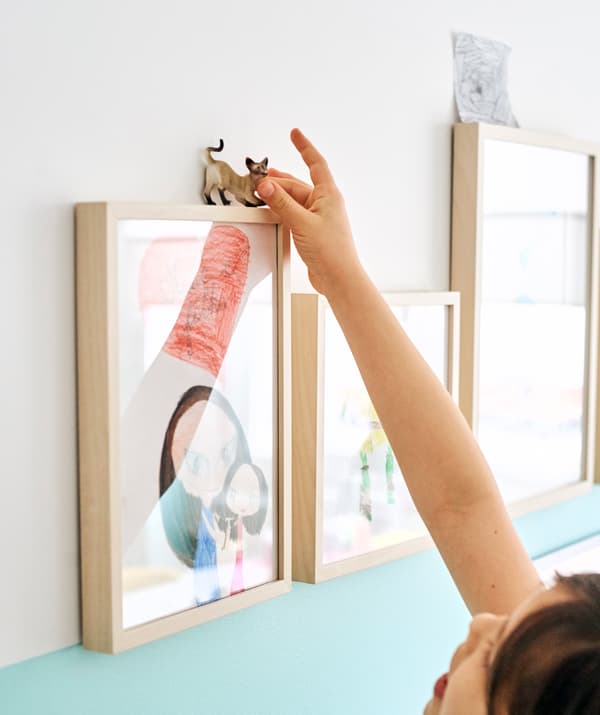 Des dessins d’enfants dans des cadres en bois alignés sur un mur. Une fillette met une figurine de chat en équilibre sur l’un d’eux.