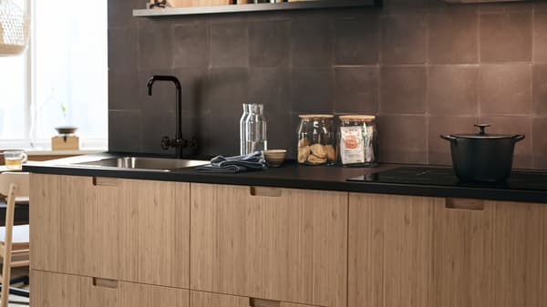 Des façades de cuisine FRÖJERED en bambou, avec divers bocaux en verre sur un comptoir et une casserole sur une plaque de cuisson.
