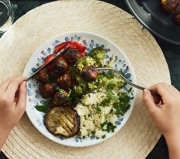 Des mains avec un couteau et une fourchette au-dessus d'une assiette végétarienne, dont des boulettes de légumes ALLEMANSRÄTTEN, posée sur un set de table.