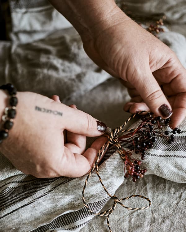 Des mains nouant un nœud décoratif fait de ficelle naturelle et de couleur noire et de baies sauvages de couleur prune autour d’une serviette de table.
