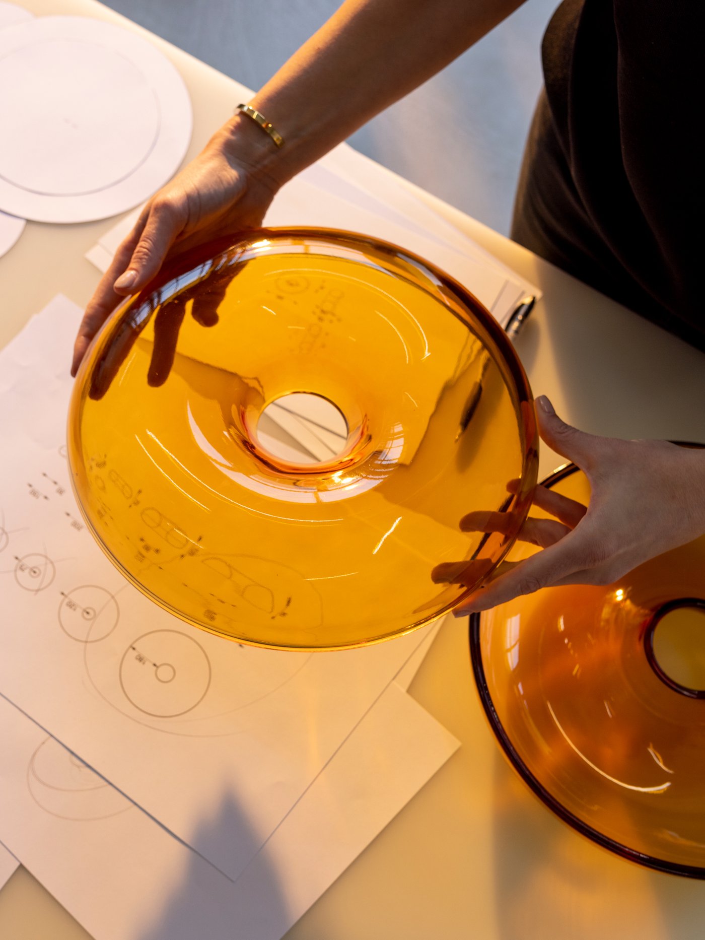 Des mains tiennent le couvercle d’un bol en forme de beigne VARMBLIXT orange au-dessus d’une table sur laquelle se trouvent des croquis de l’objet.
