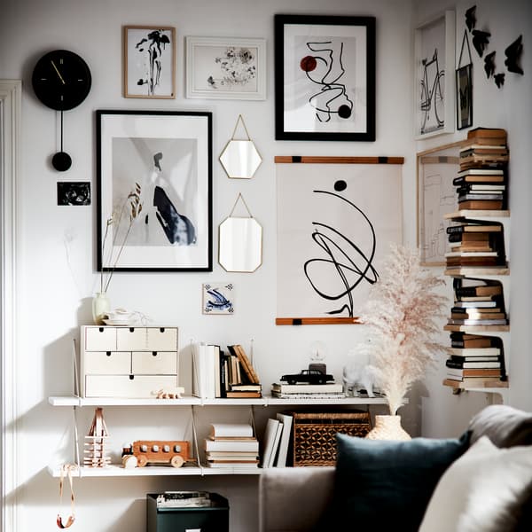 Des murs décorés de miroirs avec des bordures dorées, d’affiches encadrées de différentes tailles et couleurs, de tablettes blanches et d’une bibliothèque verticale.